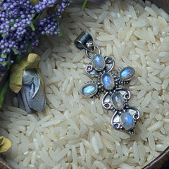 A sterling silver rainbow moonstone cross pendant placed on a bowl of rice grains, with dried flowers and purple berries in the background.