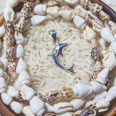A sterling silver dolphin pendant is displayed against a background of white rice and surrounded by small seashells, emphasizing its metallic shine and detailed craftsmanship.